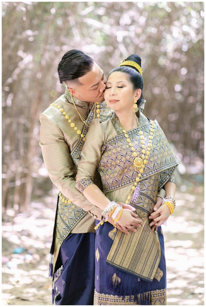 Groom in Laotian navy and gold traditional wedding attire kisses bride on cheek for Laotian wedding portrait session in tunnel of trees at North Richland HIll's park for Jenny Bui of Picture Bouquet Studio, a wedding photography studio serving the Dallas and surrounding areas. 