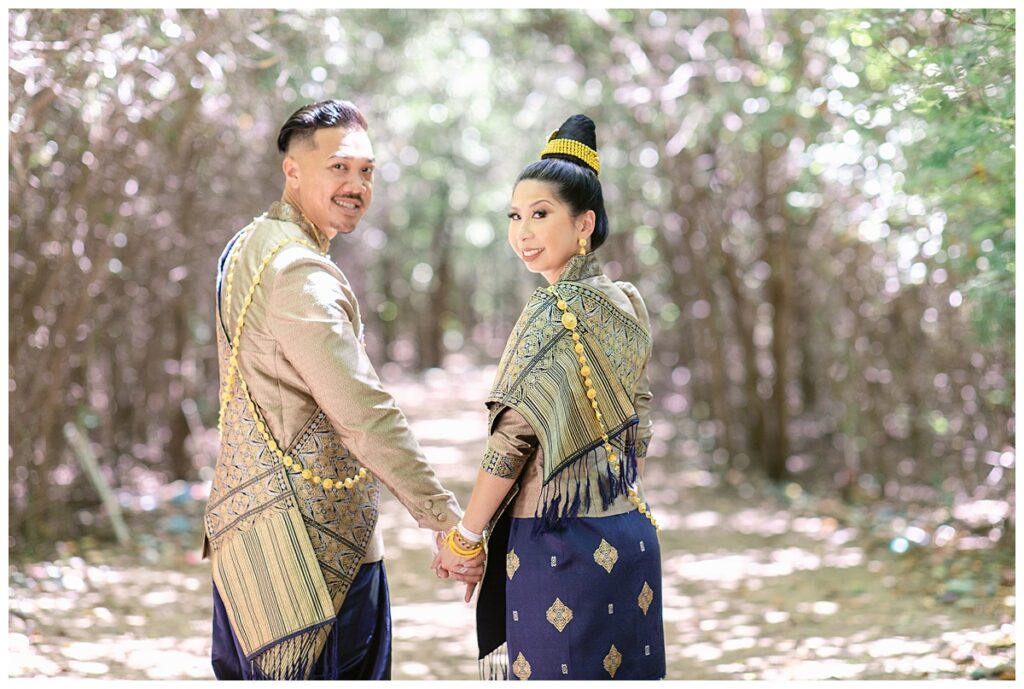 Bride and groom in Laotian navy and gold traditional wedding attire holds hands and looks back camera for Laotian wedding portrait session in tunnel of trees at North Richland HIll's park for Jenny Bui of Picture Bouquet Studio, a wedding photography studio serving the Dallas and surrounding areas. 