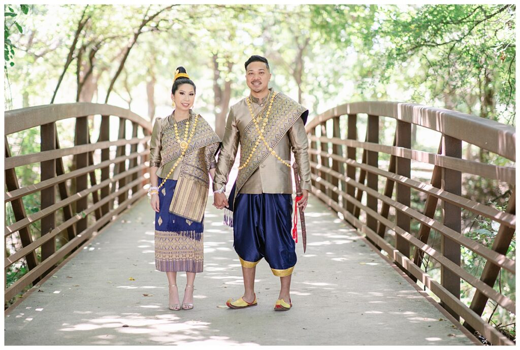 Bride and groom in Laotian navy and gold traditional wedding attire holds hands and looks at camera for Laotian wedding portrait session on bridge at North Richland HIll's park for Jenny Bui of Picture Bouquet Studio, a wedding photography studio serving the Dallas and surrounding areas. 
