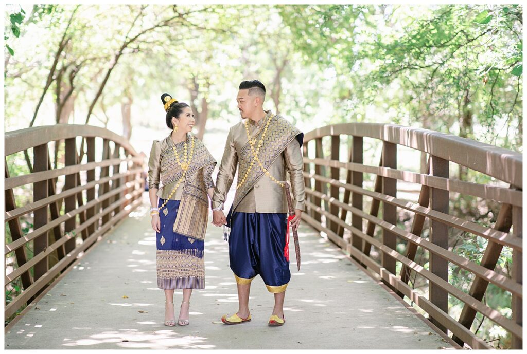 Bride and groom in Laotian navy and gold traditional wedding attire holds hands and looks at each other for Laotian wedding portrait session on bridge at North Richland HIll's park for Jenny Bui of Picture Bouquet Studio, a wedding photography studio serving the Dallas and surrounding areas. 