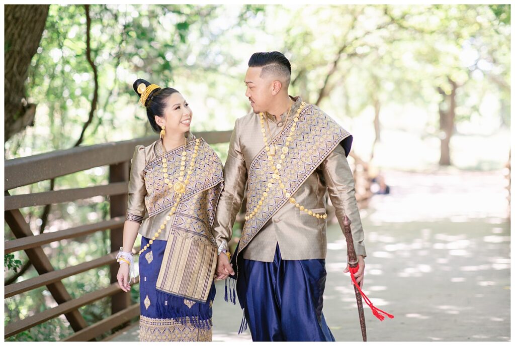Bride and groom in Laotian navy and gold traditional wedding attire holds hand while walking on bridge and looking at each other for Laotian wedding portrait session on bridge at North Richland HIll's park for Jenny Bui of Picture Bouquet Studio, a wedding photography studio serving the Dallas and surrounding areas. 