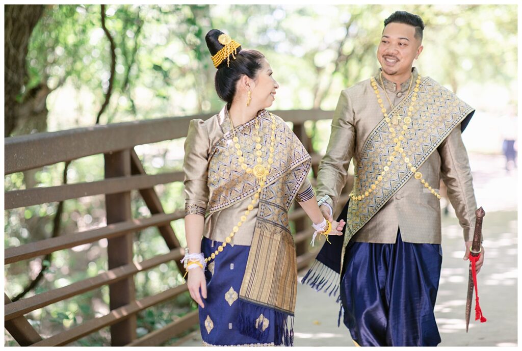 Bride in Laotian navy and gold traditional wedding attire pulls groom's hand and looks back at him on bridge for Laotian wedding portrait session on bridge at North Richland HIll's park for Jenny Bui of Picture Bouquet Studio, a wedding photography studio serving the Dallas and surrounding areas. 