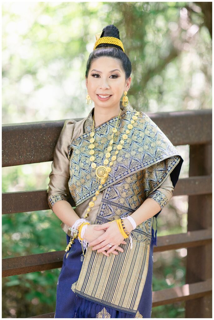 Bride in Laotian navy and gold traditional wedding attire poses with hands together at waist on bridge for Laotian wedding portrait session at North Richland HIll's park for Jenny Bui of Picture Bouquet Studio, a wedding photography studio serving the Dallas and surrounding areas. 