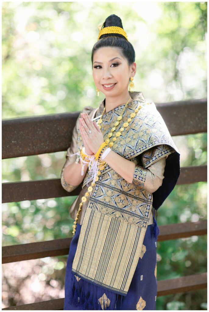 Bride in Laotian navy and gold traditional wedding attire poses  with hands together on bridge for Laotian wedding portrait session at North Richland HIll's park for Jenny Bui of Picture Bouquet Studio, a wedding photography studio serving the Dallas and surrounding areas. 