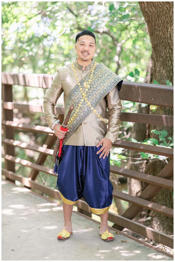 Groom in Laotian navy and gold traditional wedding attire poses on bridge for Laotian wedding portrait session at North Richland HIll's park for Jenny Bui of Picture Bouquet Studio, a wedding photography studio serving the Dallas and surrounding areas. 