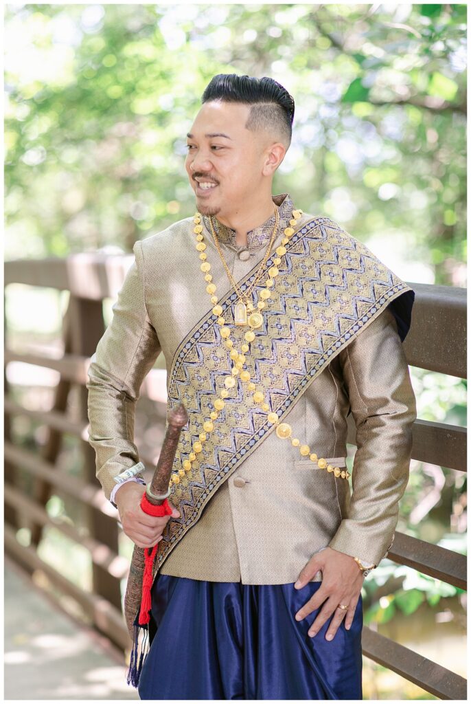 Groom in Laotian navy and gold traditional wedding attire poses with dagger on bridge for portrait session at North Richland HIll's park for Jenny Bui of Picture Bouquet Studio, a wedding photography studio serving the Dallas and surrounding areas. 