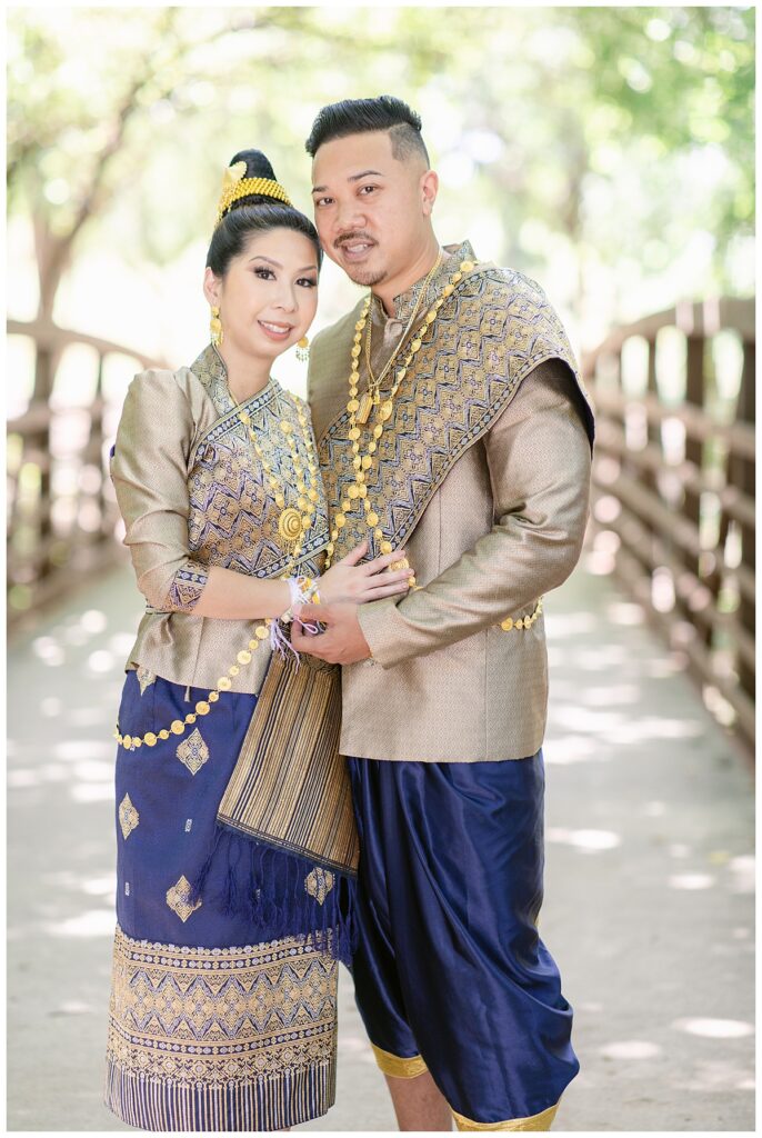 Bride and groom in Laotian navy and gold traditional wedding attire poses  with hands together and looking at camera for Laotian wedding portrait session on bridge at North Richland HIll's park for Jenny Bui of Picture Bouquet Studio, a wedding photography studio serving the Dallas and surrounding areas. 