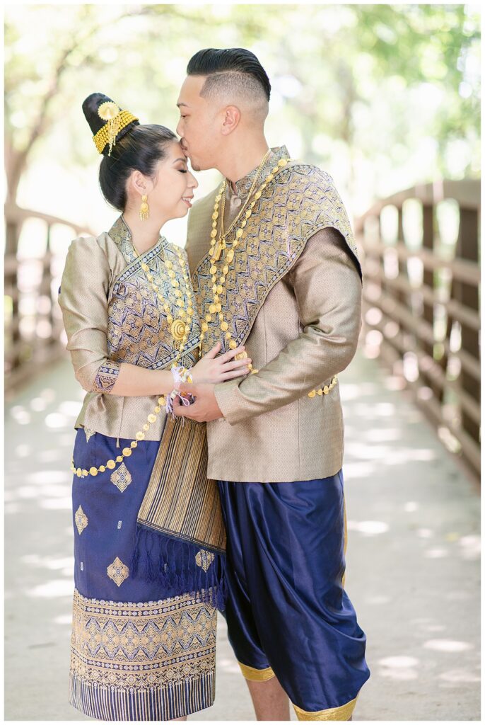 Groom in Laotian navy and gold traditional wedding attire kisses bride's forehead for Laotian wedding portrait session at North Richland HIll's park for Jenny Bui of Picture Bouquet Studio, a wedding photography studio serving the Dallas and surrounding areas. 