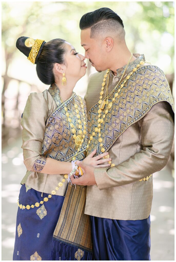 Bride and groom in Laotian navy and gold traditional wedding attire poses  with hands together at waist and faces held close together for Laotian wedding portrait session at North Richland HIll's park for Jenny Bui of Picture Bouquet Studio, a wedding photography studio serving the Dallas and surrounding areas. 