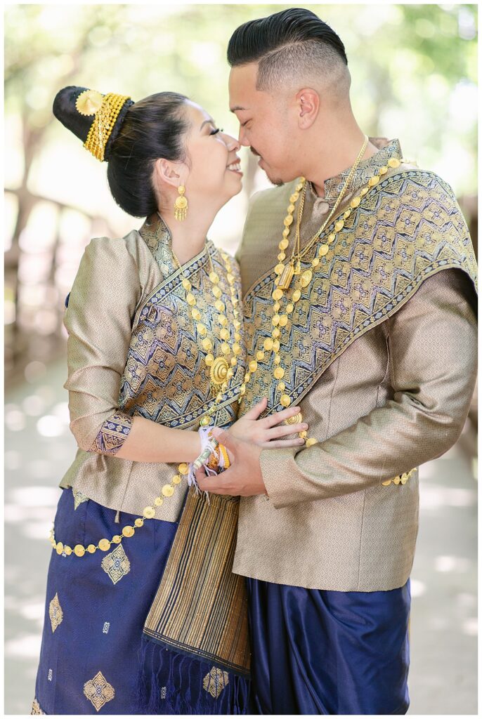 Bride and groom in Laotian navy and gold traditional wedding attire poses  with heads close together and smiling at each other for Laotian wedding portrait session at North Richland HIll's park for Jenny Bui of Picture Bouquet Studio, a wedding photography studio serving the Dallas and surrounding areas. 