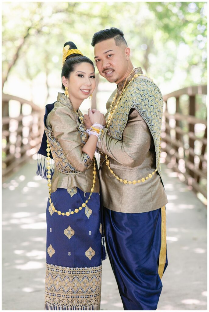 Bride and groom in Laotian navy and gold traditional wedding attire poses  with hands together and looking at camera for Laotian wedding portrait session at North Richland HIll's park for Jenny Bui of Picture Bouquet Studio, a wedding photography studio serving the Dallas and surrounding areas. 