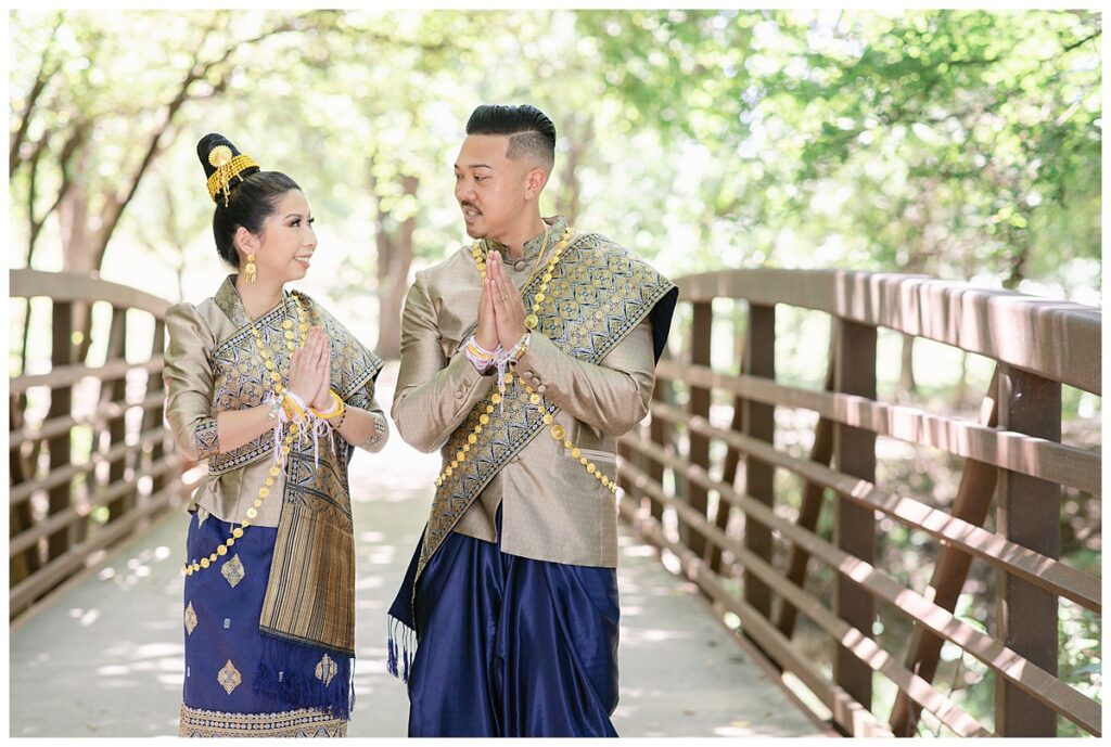 Bride and groom in Laotian navy and gold traditional wedding attire poses  with hands together and looking at each other for Laotian wedding portrait session on bridge at North Richland HIll's park for Jenny Bui of Picture Bouquet Studio, a wedding photography studio serving the Dallas and surrounding areas. 