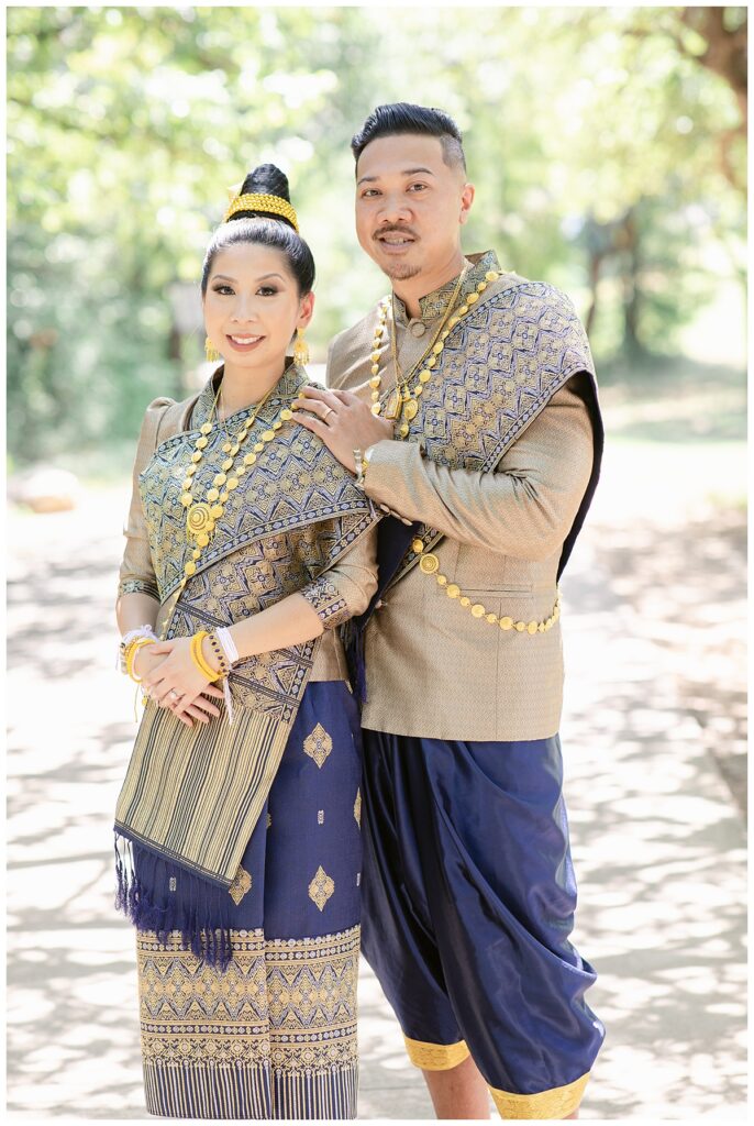 Bride and groom in Laotian navy and gold traditional wedding attire poses  along walkway with groom's hand on bride's shoulder for portrait session at North Richland HIll's park for Jenny Bui of Picture Bouquet Studio, a wedding photography studio serving the Dallas and surrounding areas. 