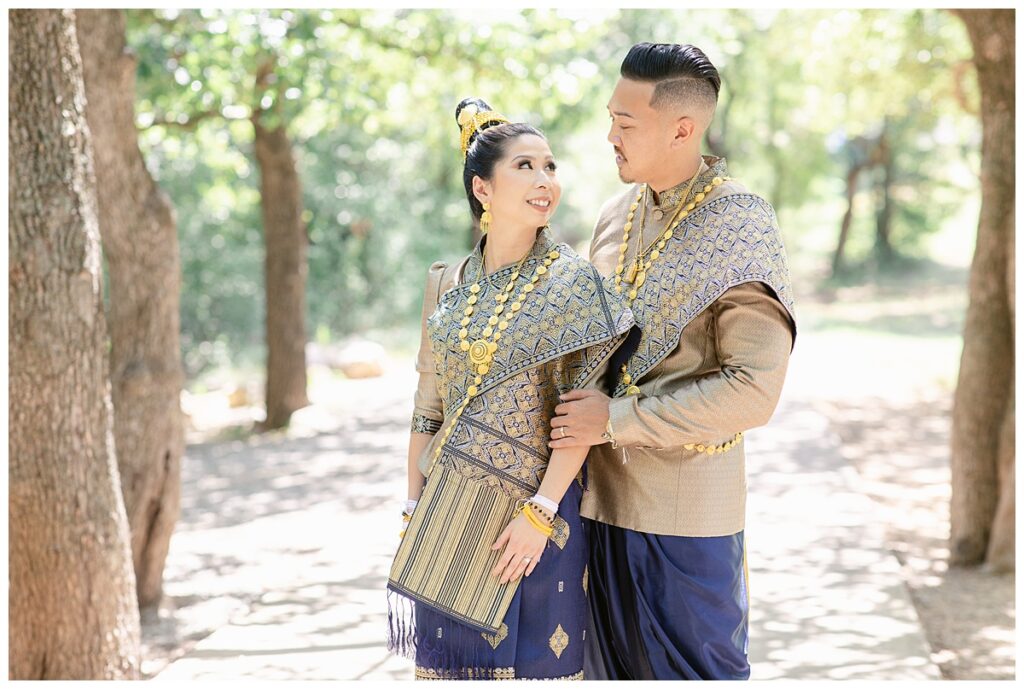 Bride and groom in Laotian navy and gold traditional wedding attire gazes at one another for portrait session at North Richland HIll's park for Jenny Bui of Picture Bouquet Studio, a wedding photography studio serving the Dallas and surrounding areas. 