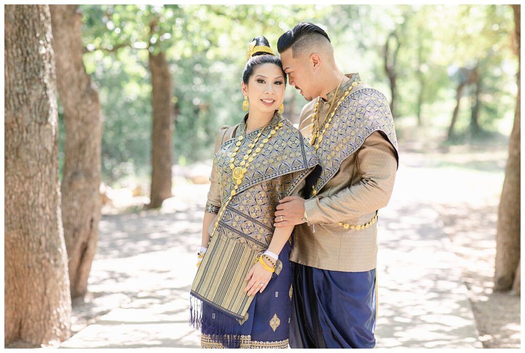 Groom in Laotian navy and gold traditional wedding attire leans into bride for portrait session at North Richland HIll's park for Jenny Bui of Picture Bouquet Studio, a wedding photography studio serving the Dallas and surrounding areas. 
