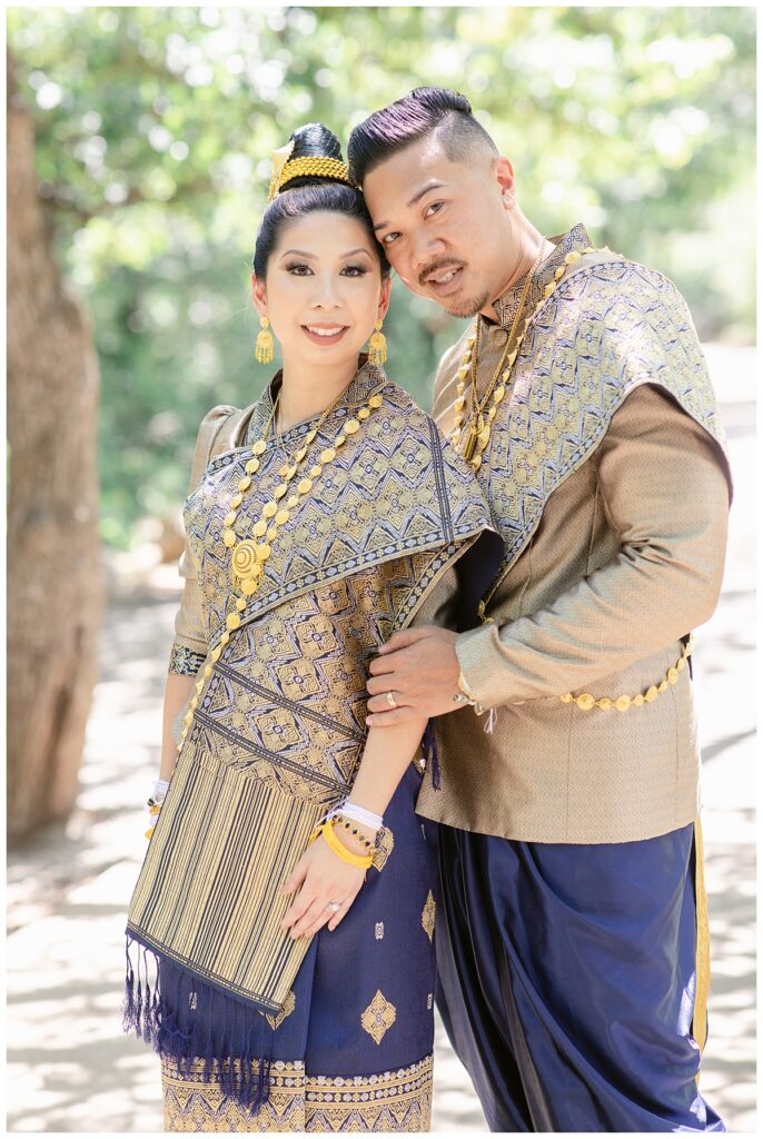 Groom in Laotian navy and gold traditional wedding attire leans into bride for portrait session at North Richland HIll's park for Jenny Bui of Picture Bouquet Studio, a wedding photography studio serving the Dallas and surrounding areas. 