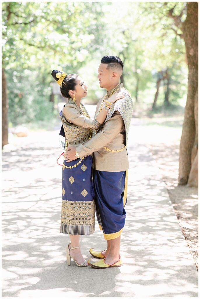 Bride and groom in Laotian navy and gold traditional wedding attire poses for portrait session with bride's hand on groom's shoulder and groom's hands on bride's waist for full body shot at North Richland HIll's park for Jenny Bui of Picture Bouquet Studio, a wedding photography studio serving the Dallas and surrounding areas. 