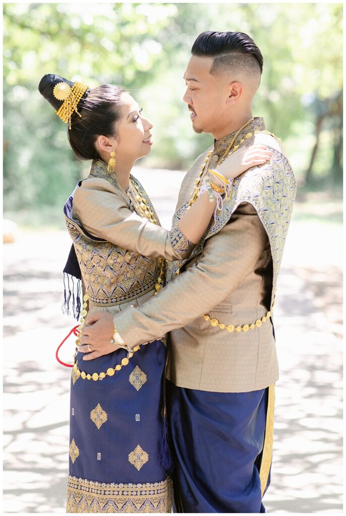 Bride and groom in Laotian navy and gold traditional wedding attire gazes at one another with bride's hand on groom's shoulders and groom's hand around bride's waist for portrait session at North Richland HIll's park for Jenny Bui of Picture Bouquet Studio, a wedding photography studio serving the Dallas and surrounding areas. 
