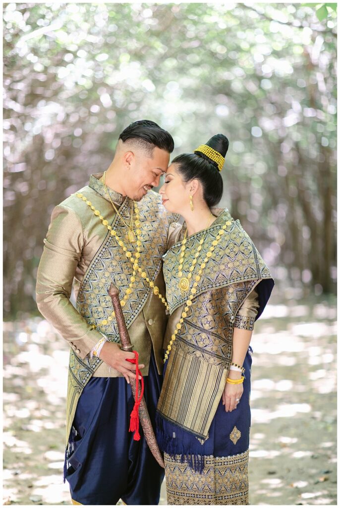 Bride and groom in Laotian navy and gold traditional wedding attire presses foreheads together gently for Laotian wedding portrait session in tunnel of trees at North Richland HIll's park for Jenny Bui of Picture Bouquet Studio, a wedding photography studio serving the Dallas and surrounding areas. 