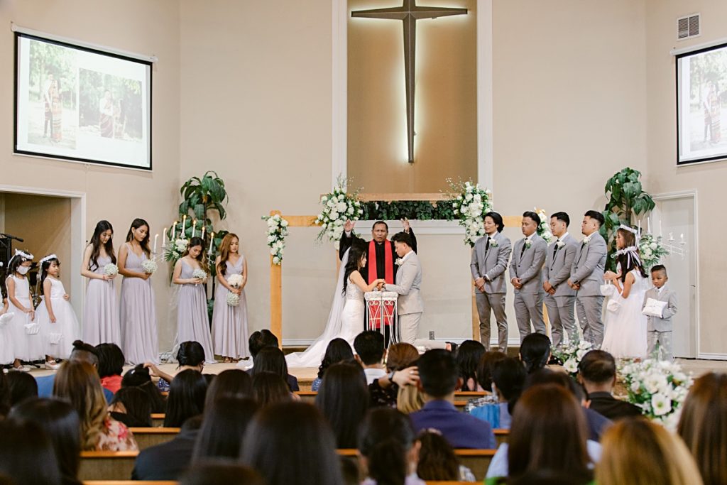 Bride and groom kneeling during wedding ceremony with bridal party on stage photographed by Picture Bouquet Studio at Chin Revival Church. 