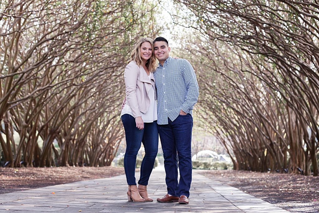Young engaged couple poses under tree walkway at the Dallas Arboretum photographed by Dallas wedding photographer Jenny Bui of Picture Bouquet Studio. 