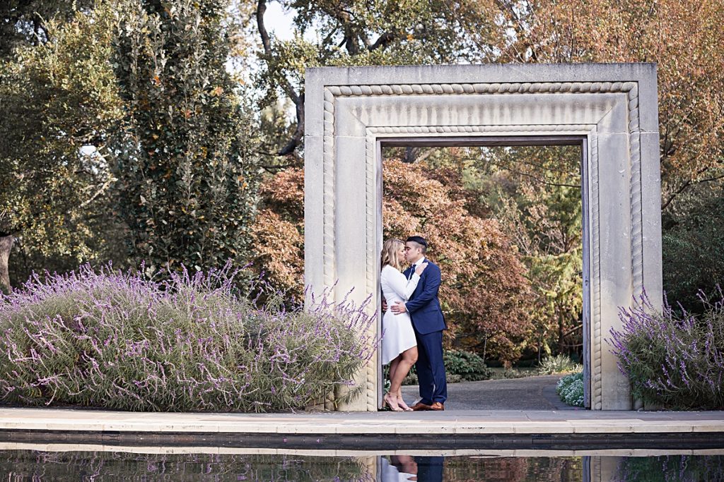 Young man in navy suit kisses fiancee in long sleeve white dress under the photo frame at the Dallas Arboretum for their engagement session photographed by Dallas wedding photographer Jenny Bui of Picture Bouquet Studio. 