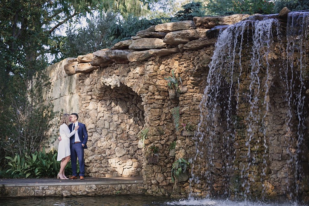 Young man in navy suit kisses fiancee in long sleeve white dress next to the waterfall at the Dallas Arboretum for their engagement session photographed by Dallas wedding photographer Jenny Bui of Picture Bouquet Studio. 
