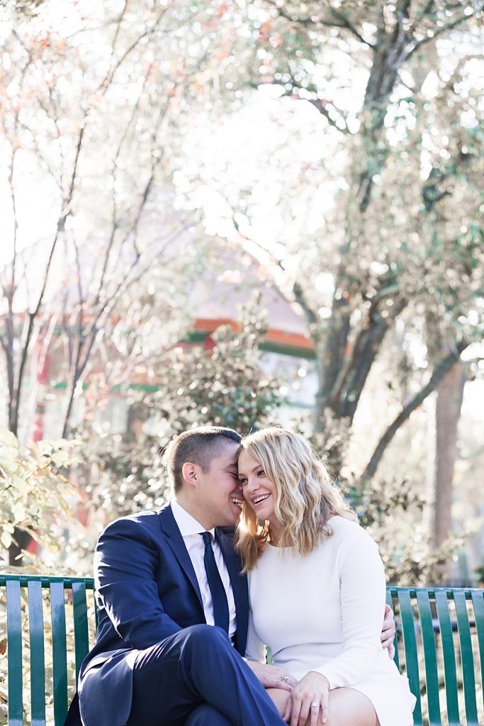 Young man in navy suit leans in and whispers in fiancee in long sleeve white dress at the Dallas Arboretum for their engagement session photographed by Dallas wedding photographer Jenny Bui of Picture Bouquet Studio. 