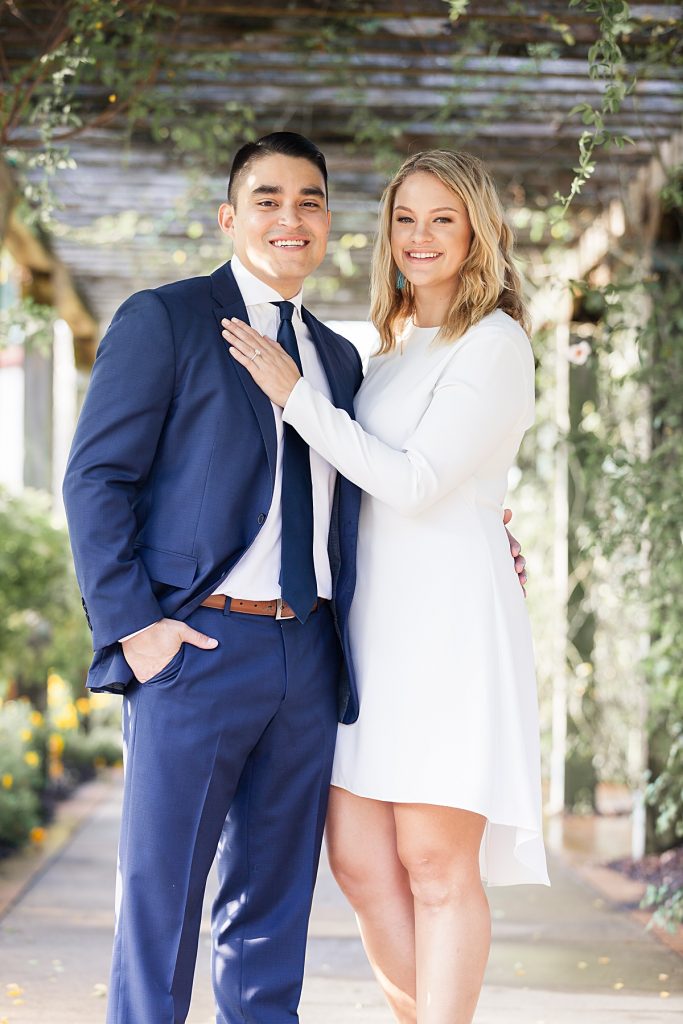 Young man in navy suit poses with fiancee in long sleeve white dress under gazebo walkway at the Dallas Arboretum for their engagement session photographed by Dallas wedding photographer Jenny Bui of Picture Bouquet Studio. 