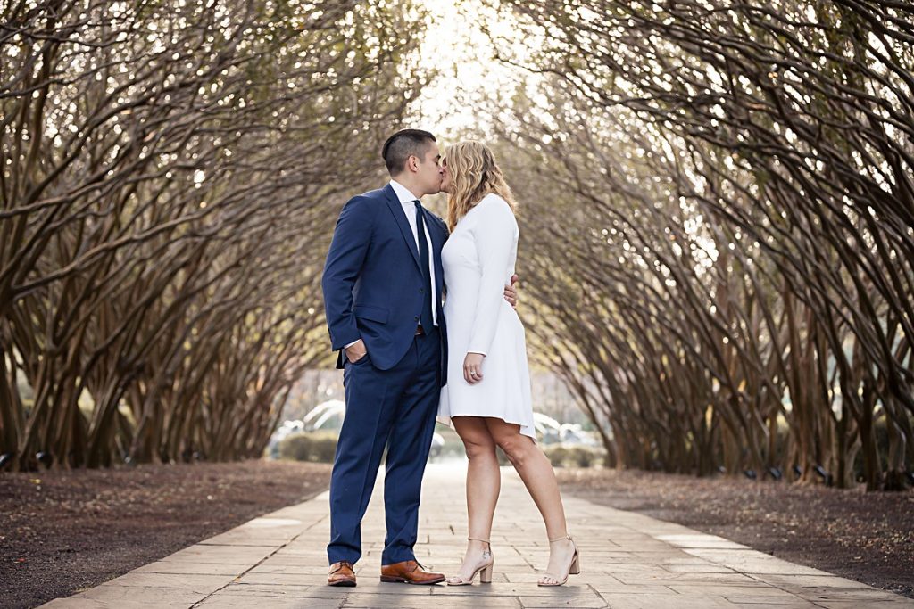 Dallas engaged couple kisses under the tree walkway at the Dallas Arboretum for their engagement session photographed by Dallas wedding photographer Jenny Bui of Picture Bouquet Studio. 