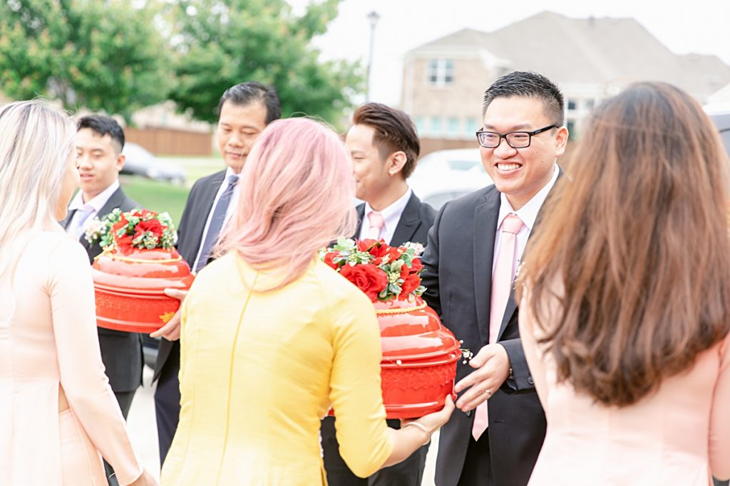 Vietnamese groomsmen handing off red trays of gifts to bridesmaids in ao dai for traditional Vietnamese tea ceremony photographed by Dallas Vietnamese wedding photographer Jenny Bui of Picture Bouquet Studio. 