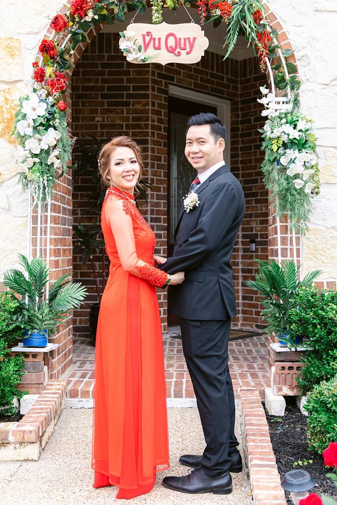 Vietnamese bride in red ao dai holds groom in black suit's hands as they pose for the camera for traditional Vietnamese tea ceremony photographed by Dallas Vietnamese wedding photographer Jenny Bui of Picture Bouquet Studio. 
