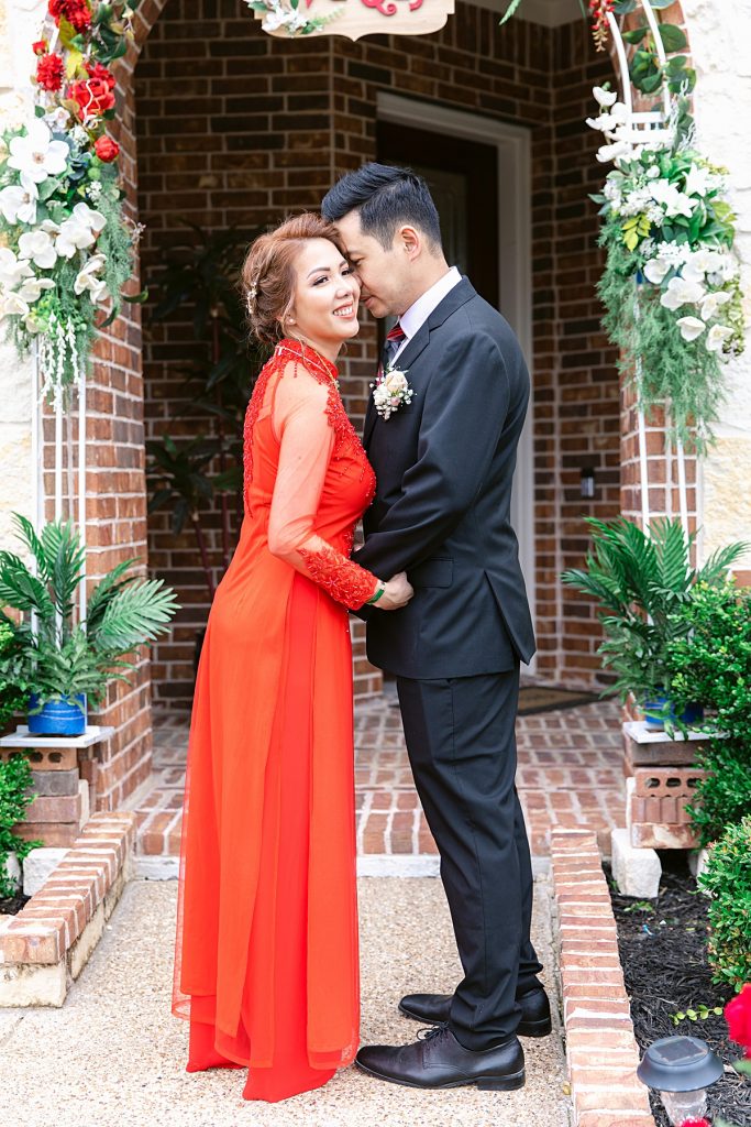 Vietnamese bride in red ao dai and groom in black suit poses in front of bride's home for traditional Vietnamese tea ceremony photographed by Dallas Vietnamese wedding photographer Jenny Bui of Picture Bouquet Studio. 