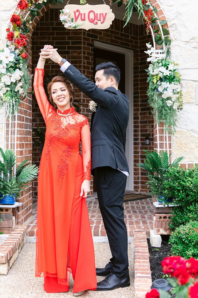 Vietnamese groom in black suit twirls bride in red ao dai around in front of Vu Quy sign for traditional Vietnamese tea ceremony photographed by Dallas Vietnamese wedding photographer Jenny Bui of Picture Bouquet Studio. 