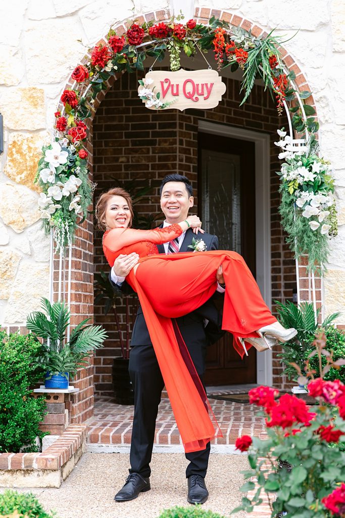 Groom in black suit carries bride in red ao dai in front of Vu Quy sign Vietnamese groom in black suit twirls bride in red ao dai around for traditional Vietnamese tea ceremony photographed by Dallas Vietnamese wedding photographer Jenny Bui of Picture Bouquet Studio. 