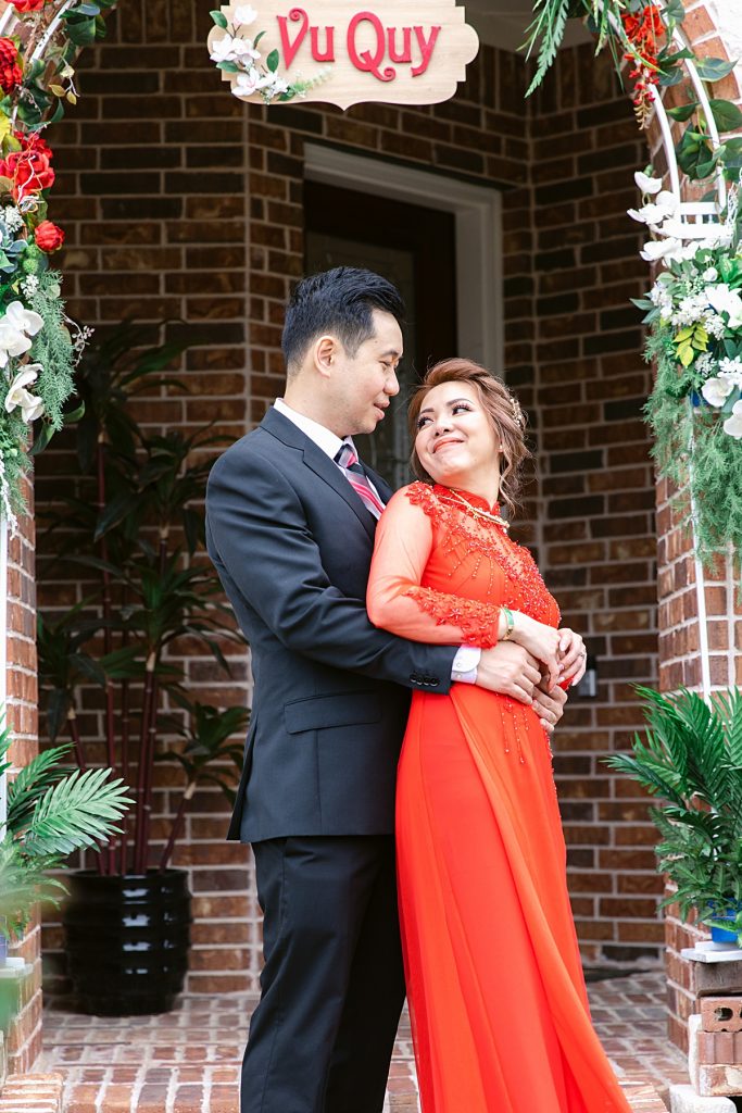 Vietnamese bride in red ao ai and groom in black suit poses in front of Vu Quy sign for traditional Vietnamese tea ceremony photographed by Dallas Vietnamese wedding photographer Jenny Bui of Picture Bouquet Studio. 