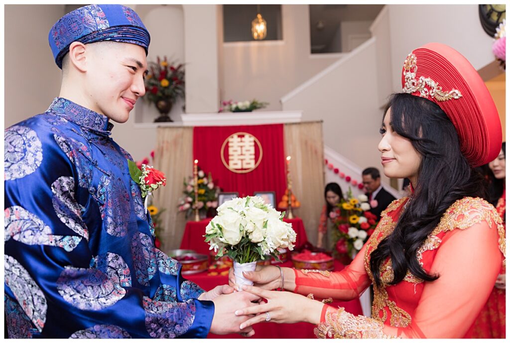 Groom in blue ao dai hands bride in red ao dai bouquet during traditional Vietnamese tea ceremony photographed by Jenny Bui of Picture Bouquet Studio. 