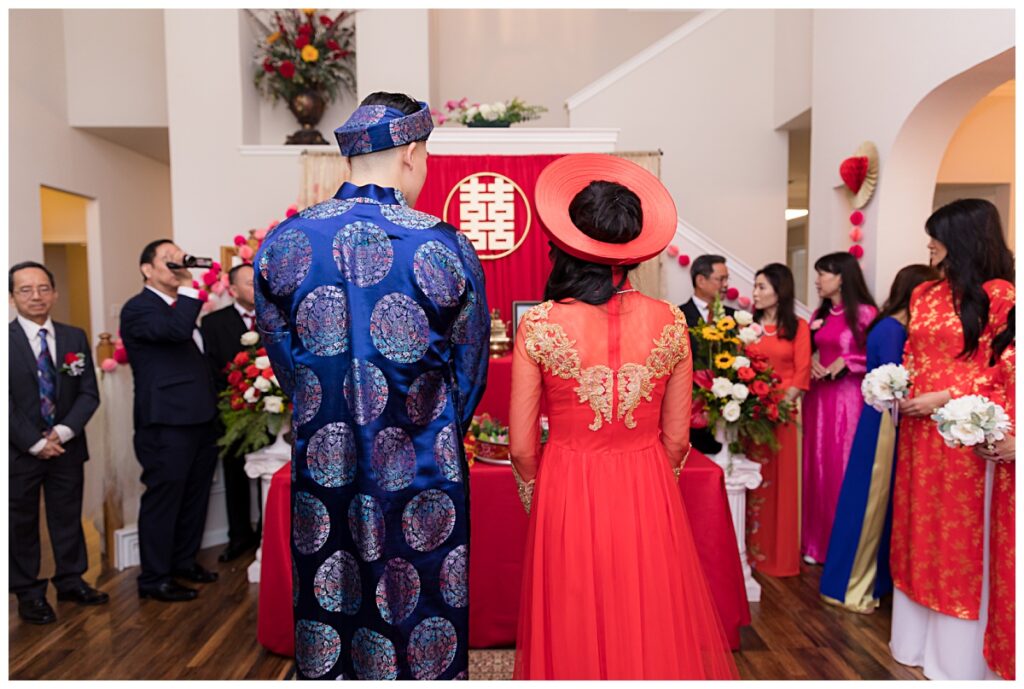 Groom in blue ao dai and bride in red ao dai stands in front of altar during Vietnamese tea ceremony photographed by Jenny Bui of Picture Bouquet Studio. 