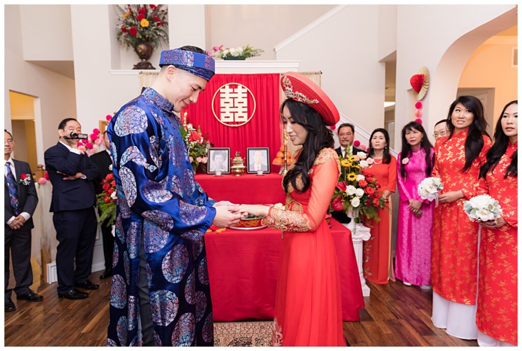 Groom in blue ao dai places ring on bride's finger during traditional Vietnamese tea ceremony photographed by Jenny Bui of Picture Bouquet Studio. 