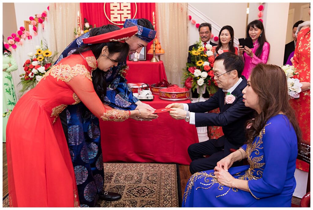 Bride and groom in red and blue ao dai presenting tea and receiving red envelopes from bride's parents during traditional Vietnamese tea ceremony photographed by Jenny Bui of Picture Bouquet Studio. 