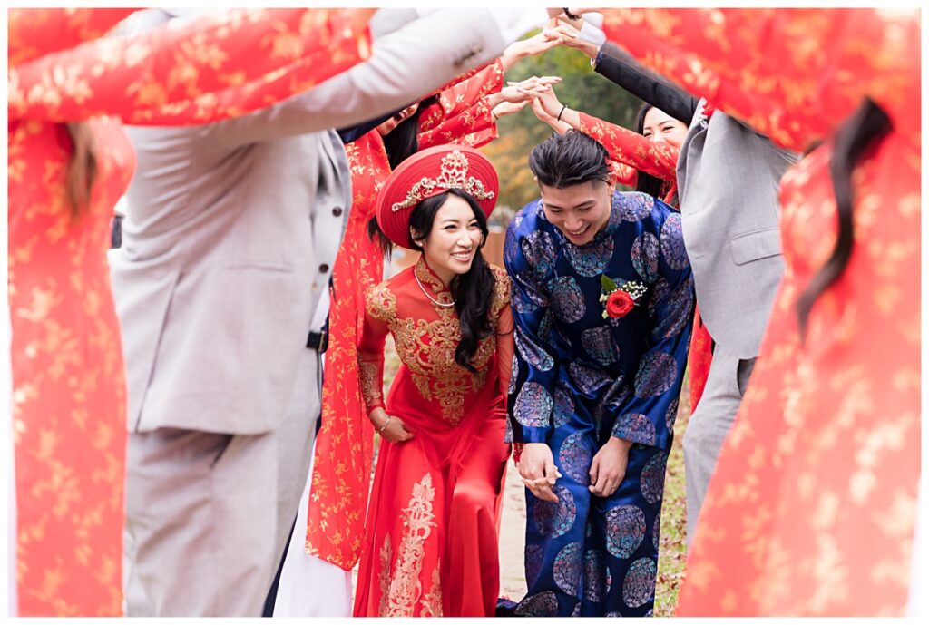 Bride and groom in red and blue ao dai goes under tunnel of bridesmaid and groomsmen arms during Dallas Vietnamese tea ceremony photographed by Jenny Bui of Picture Bouquet Studio. 