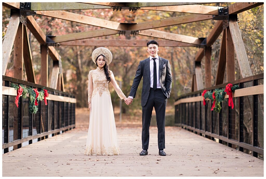 Bride in yellow ao dai and groom in grey suit poses looking at camera on bridge during Prairie creek portrait session photographed by Dallas wedding photographer Jenny Bui of Picture Bouquet Studio. 