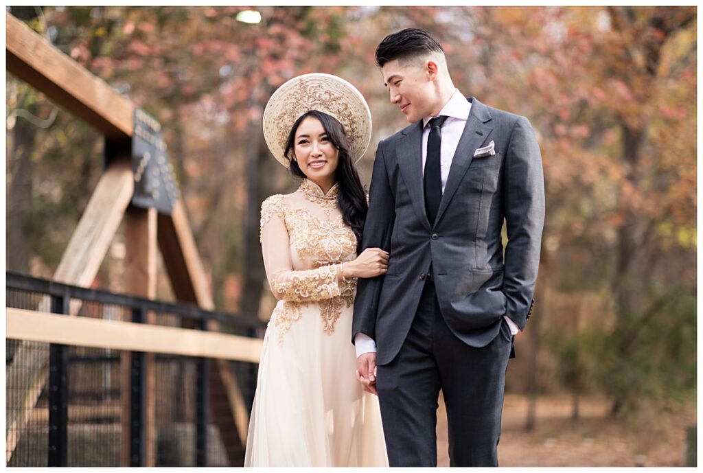 Vietnamese bride in gorgeous ao dai and groom in grey suit walks hand in hand on bridge during Prairie creek portrait session photographed by Dallas wedding photographer Jenny Bui of Picture Bouquet Studio. 