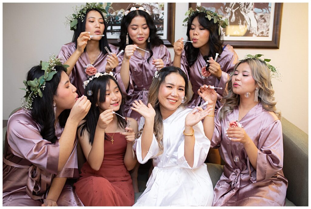 Bridesmaids in lavender robes blow bubbles at bride in white robe during getting ready portion of The Pearl wedding in Arlington, TX photographed by Picture Bouquet Studio. 