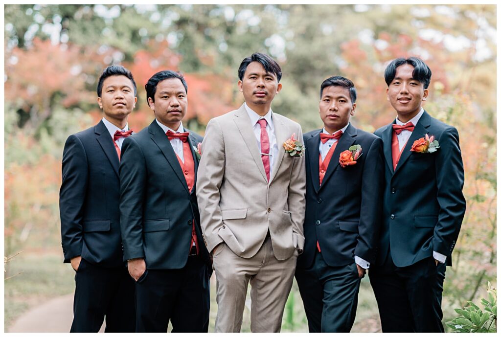 Groomsmen in burnt orange vest and bowtie poses with groom in burnt orange tie and tan suit at Fort Worth Japanese Garden photographed by Jenny Bui of Picture Bouquet Studio. 