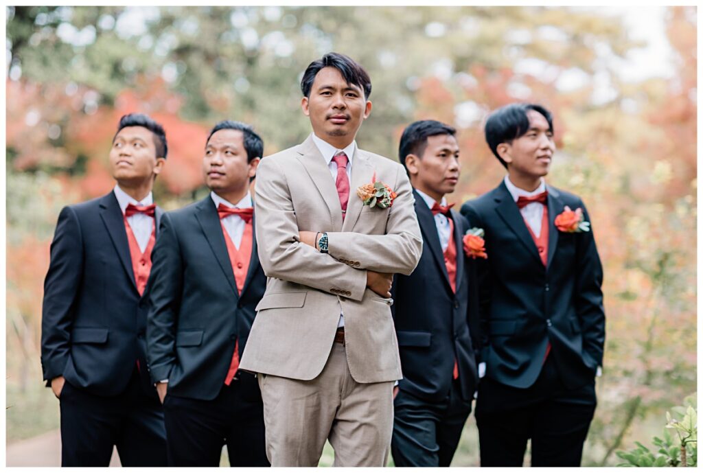 Groomsmen in burnt orange vest and bowtie poses with groom in tan suit and burnt orange tie at Fort Worth Japanese Garden photographed by Jenny Bui of Picture Bouquet Studio. 