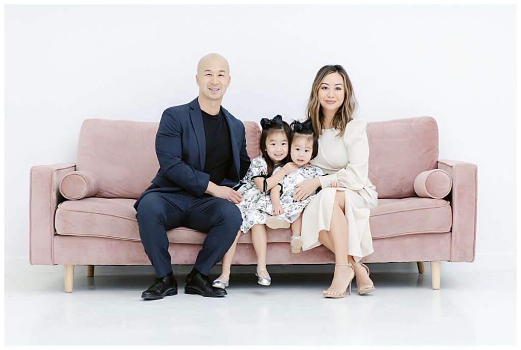 Family of four in dressy attire posing on light pink sofa for a family session at The Lumen Room photographed by Picture Bouquet Studio. 