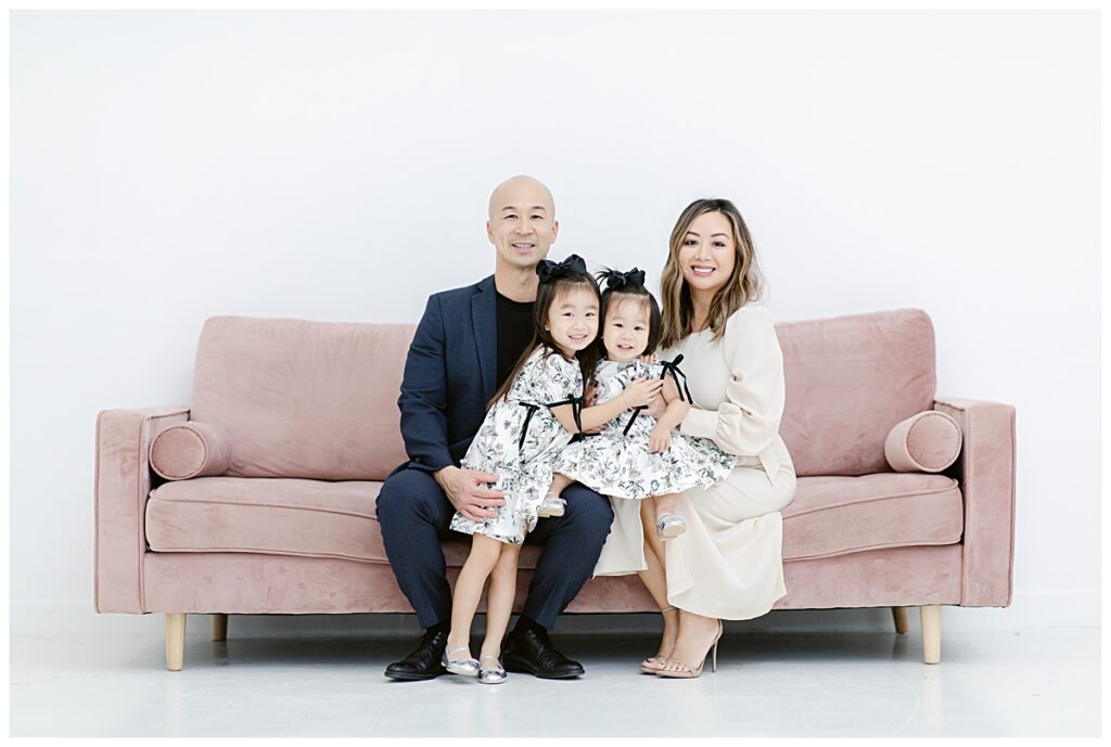 Family of four smiling at camera sitting on light pink sofa during The Lumen Room family session photographed by Jenny Bui of Picture Bouquet Studio. 