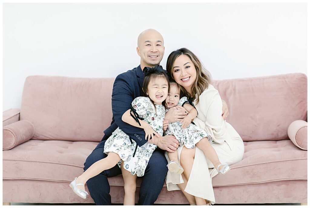 Family of four hugging in dressy attire sitting on light pink sofa during The Lumen Room family session photographed by Dallas family photographer Jenny Bui of Picture Bouquet Studio. 