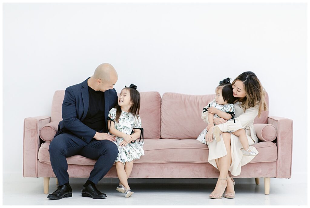 Fun family pose on light pink sofa of daddy and older daughter on one end and mommy and younger daughter on opposite end photographed at the Lumen Room in Plano by Picture Bouquet Studio. 